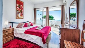 Een slaapkamer met een bed, een kaptafel, een nachtkastje, een rood tapijt, een schilderij en uitzicht op buiten.