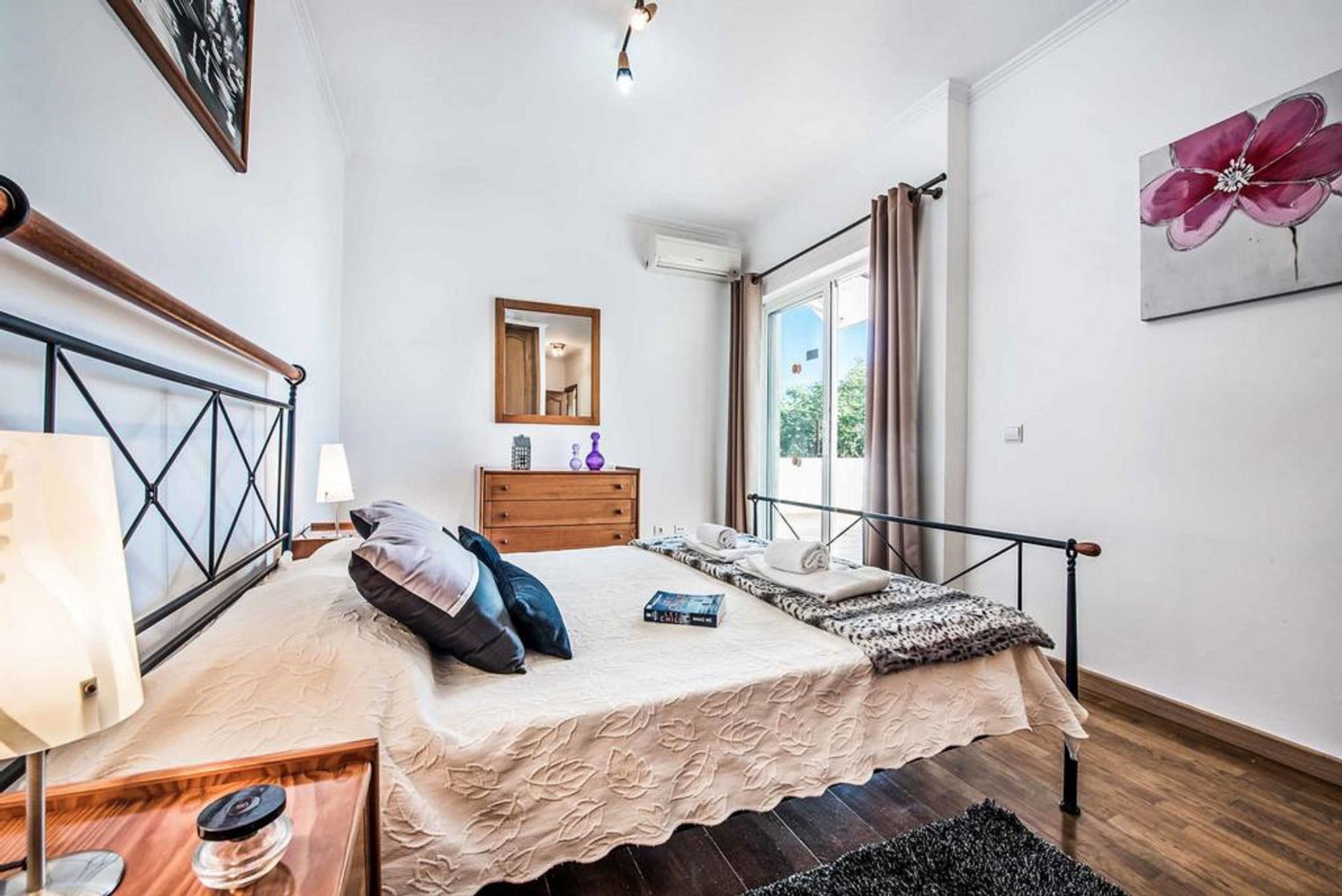 Une chambre à coucher comprenant un lit, une table de chevet, une lampe, une commode, un miroir et une fenêtre avec des rideaux.