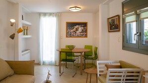 A modern living room with a sofa, a dining table with chairs, a small table with a plant, and a window with blinds. - Eclectic Urban Nest in Heraklion (Heraklion)