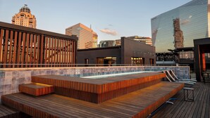 Zona de piscina en la azotea con escalones de madera y sillones de descanso, rodeada de edificios modernos y un cielo despejado.