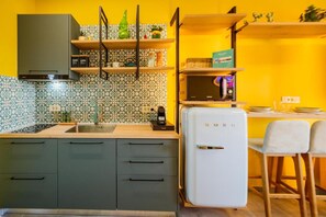 Una cocina compacta con microondas, refrigerador y lavamanos. Hay estantes de madera sobre la encimera y una pequeña mesa con sillas.