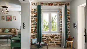 A living room with a stone wall, a window with curtains, a sofa, a coffee table, and a small side table with a picture frame. - The Coastal Rock House (Volos)