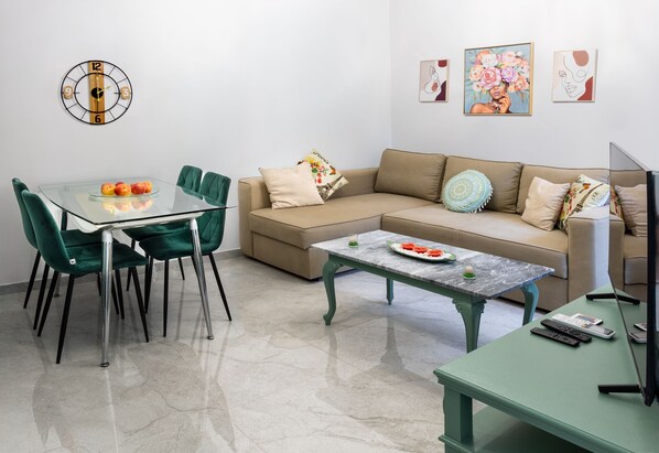A modern living room with a glass dining table, green chairs, a beige sofa, a green coffee table, and a TV on a green stand.