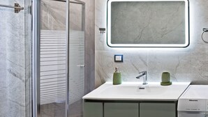 A modern bathroom with a glass shower enclosure, a sink with a mirror, and a washing machine. - The Coastal Rock House (Volos)