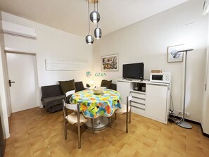 A small, well-lit room with a dining table, chairs, a sofa, a television, a microwave, and a floor lamp. - 3 rooms 4 beds Conchiglia area. Il Gabbiano-Rif. Il gabbiano (San Vincenzo)