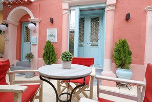 Un bâtiment rose avec une porte bleue, une petite table sur laquelle est posée une plante en pot, et des chaises rouges.