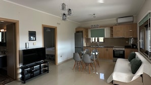 En moderne stue med et fladskærms-tv, et glasbord og en sofa. Der er et spiseområde med et bord og stole. Køkkenet har træskabe og en indbygget ovn.