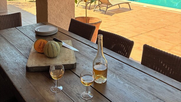 A wooden table with a melon, a bottle, and glasses, overlooking a swimming pool and lush greenery. - 4-bedroom villa with private pétanque pool and padel court (Le Muy)