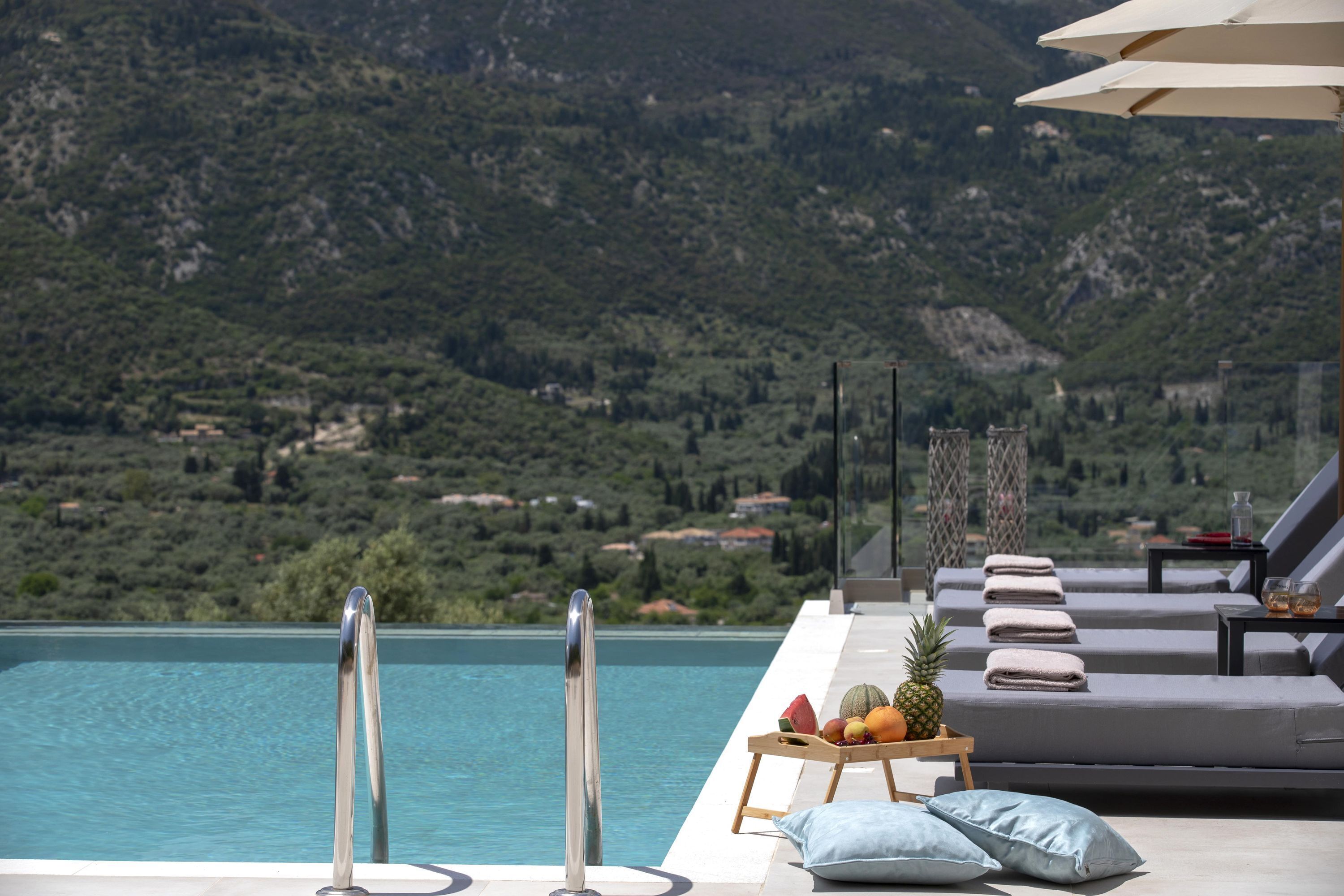 A poolside area with a view of mountains, a lounge with cushions, and a fruit tray.