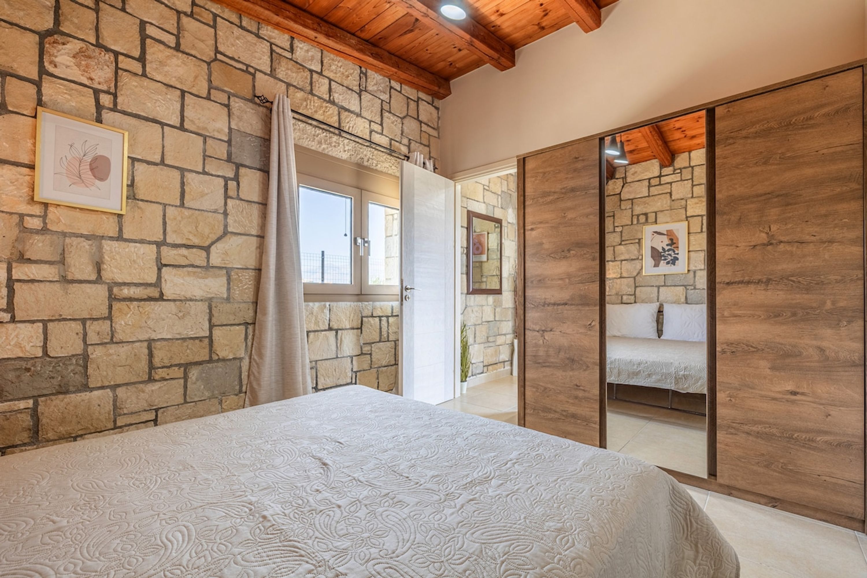 Un dormitorio con pared de piedra, cama con colcha estampada, techo de madera y un espejo grande.