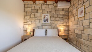 A bedroom with a stone wall, a bed with white linens, two bedside lamps, and framed artwork on the wall. - Elea Home by cretanhouses (Gournes)