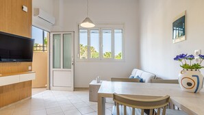 A modern kitchen with a TV, wooden paneling, a dining area with a table and chairs, a window with a view, and a vase with flowers. - The Walls Apartments | 1 (Iraklio)