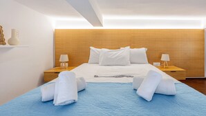 A modern hotel room with a wooden headboard, a large bed with white linens, two bedside tables with lamps, and a blue bedspread. - The Walls Apartments | 1 (Iraklio)