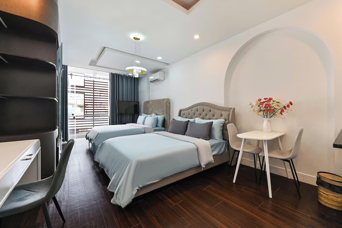 A modern hotel room with two beds, a desk, a chair, a small table, and a decorative vase.