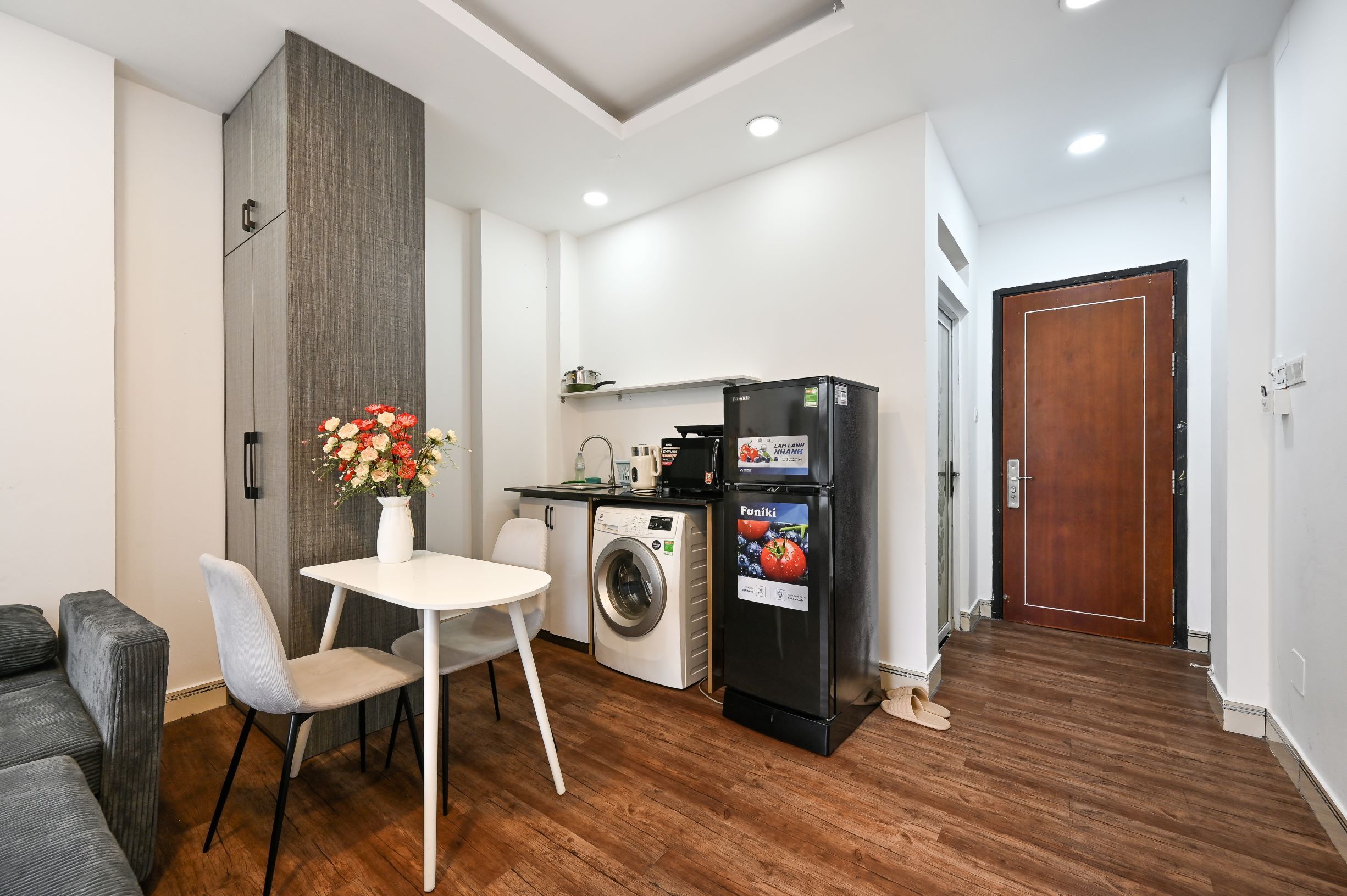 A compact kitchen with a washing machine, a refrigerator, a dining table with chairs, and a wooden floor.