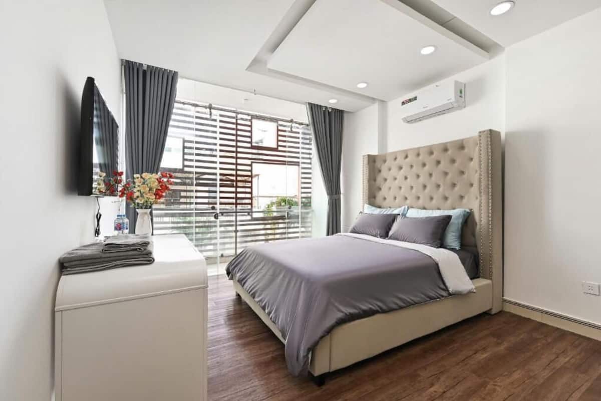 A modern bedroom with a large bed, a bedside table, a TV, and a window with blinds.