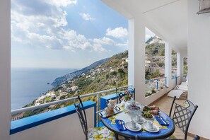 A balcony with a table set for dining, overlooking a coastal landscape with buildings and a view of the sea. - At Nonno Aurelio's: Sea View, Wi-Fi & Air Conditioning (Furore)
