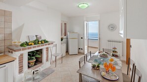 Cozinha moderna com lareira de tijolos, mesa de jantar com cadeiras e vista para o mar por uma porta aberta.