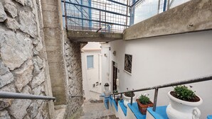 Uma escadaria de pedra estreita com corrimão de metal, que leva a um edifício branco com porta azul e uma pequena janela.