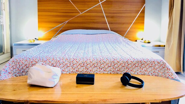 Una cama bien hecha con edredón estampado con motivos florales, una mesita de noche con una lámpara, una regleta y una cajita.