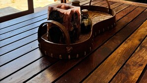 A wooden table with a woven basket containing a bottle and a container, a fireplace, and a view of greenery through large windows.