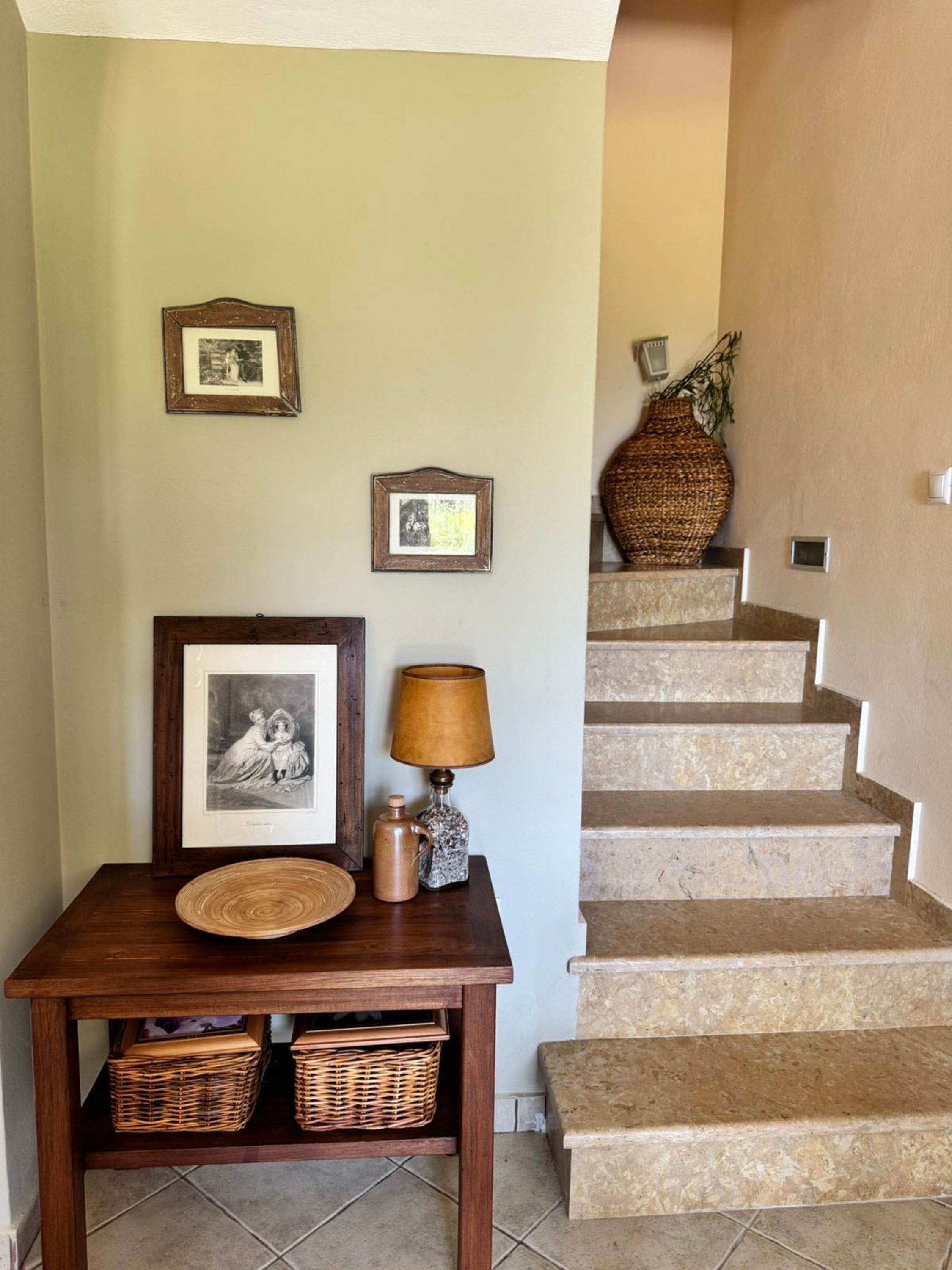 Una escalera con un muro color crema y una mesa de madera con una lámpara e imágenes enmarcadas.