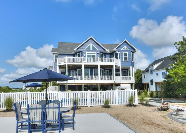 A large two-story house with multiple balconies, a patio with chairs and an umbrella, and a white picket fence.