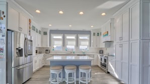 A modern kitchen with white cabinetry, a central island with stools, stainless steel appliances, and a view through the windows. - Bayfront home with private pool on Slaughter Beach! (Milford)