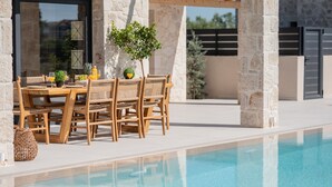 Zona junto a la piscina con una mesa y sillas de madera, rodeada de pilares de piedra y una piscina cristalina.