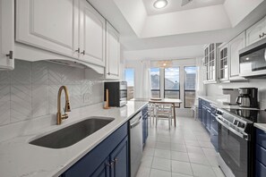 A modern kitchen with white cabinetry, a large island with a sink, and a view of the outdoors through large windows. - Ocean View, Central Island - 134 Captain John Fripp (Fripp Island)