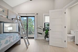 Quarto com cama beliche, escada, mesa de cabeceira com abajur, janela grande com cortinas e vista para vegetação lá fora.