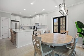 A modern kitchen with white cabinets, a central island, and a dining area with a wooden table and chairs. - Golf Course Front, Golf Cart Included, Pet Friendly - 155 Davis Love (Fripp Island)
