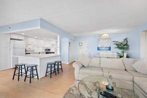 Ein modernes Wohnzimmer mit einer Couch, einem Couchtisch und einer Küchenzeile mit weißen Schränken und einem Barhocker.