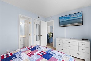 Une chambre à coucher avec un lit, une commode, un tableau représentant une plage et une porte donnant sur une autre pièce.