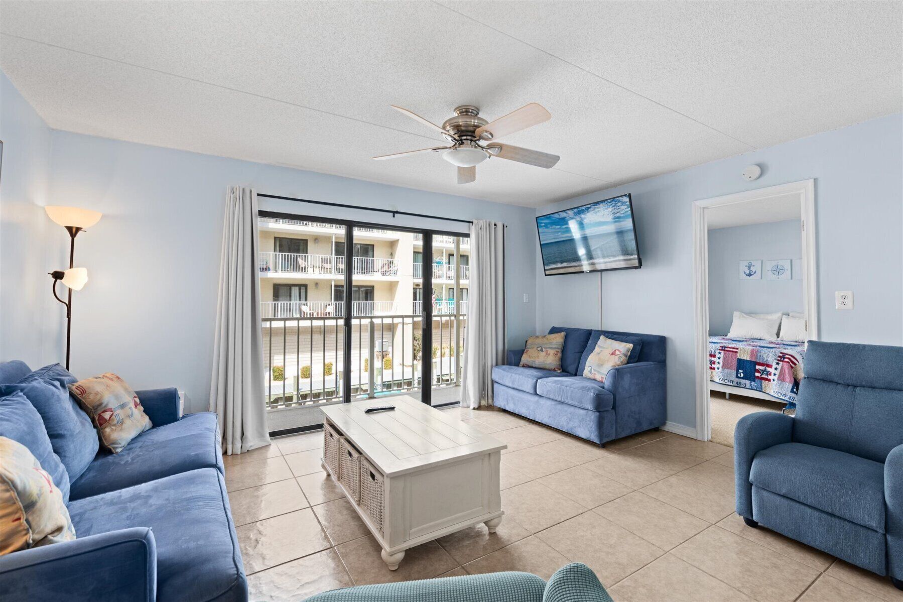 A living room with a ceiling fan, a TV mounted on the wall, and a view of the outside balcony.