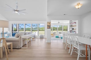 A spacious living area with a sofa, dining table, and chairs, large windows with a view of greenery, and a ceiling fan. - Gorgeous Sunsets and Marsh Views, Pet Friendly, Golf Cart Included (Fripp Island)