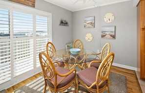 A dining area with a round glass table surrounded by wicker chairs, a view of the outdoors through large windows, and wall-mounted clocks.