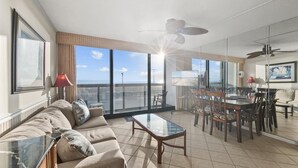 Un espacio habitable amplio con un gran ventanal que da a un balcón, un comedor con sillas y una mesa, y un ventilador de techo.