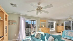 A spacious room with a teal sofa, a wooden cabinet, a ceiling fan, and a large window with a view. - Harbor View by Seaside Vacations & Sales (Ocean City)