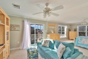 A spacious room with a teal sofa, a wooden cabinet, a ceiling fan, and a large window with a view. - Harbor View by Seaside Vacations & Sales (Ocean City)
