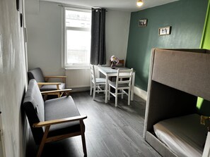 Ein kleines Zimmer mit einem Etagenbett, einem Tisch mit Stühlen, einem Heizkörper und einem Vorhangfenster.