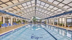 Ein Hallenbad mit Glasdecke, blauen Liegestühlen und Pflanzen rund um den Poolbereich.
