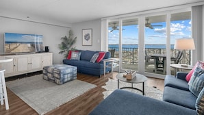 Ein Wohnzimmer mit einer Couch, einem Hocker und einem Couchtisch, einem Fernseher und Blick auf das Meer durch große Fenster.