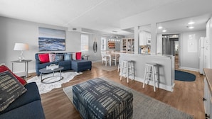 Ein modernes Wohnzimmer mit Sofa, Hocker und Couchtisch. Es gibt eine Küchenzeile mit Bar und Barhocker.