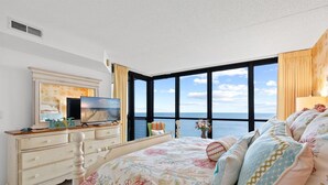 Une chambre avec une grande fenêtre, un lit recouvert d’une literie à motifs floraux, une commode et un téléviseur posé sur un meuble.