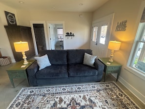 Un salon avec un canapé gris foncé, deux lampes assorties, un tapis à motifs et une porte sur laquelle est apposée une plaque indiquant « Résidence au bord du lac ».