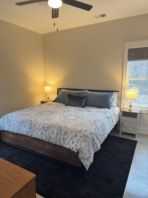 Une chambre à coucher avec un lit, un ventilateur de plafond, une table de chevet avec une lampe et une fenêtre donnant sur des arbres.
