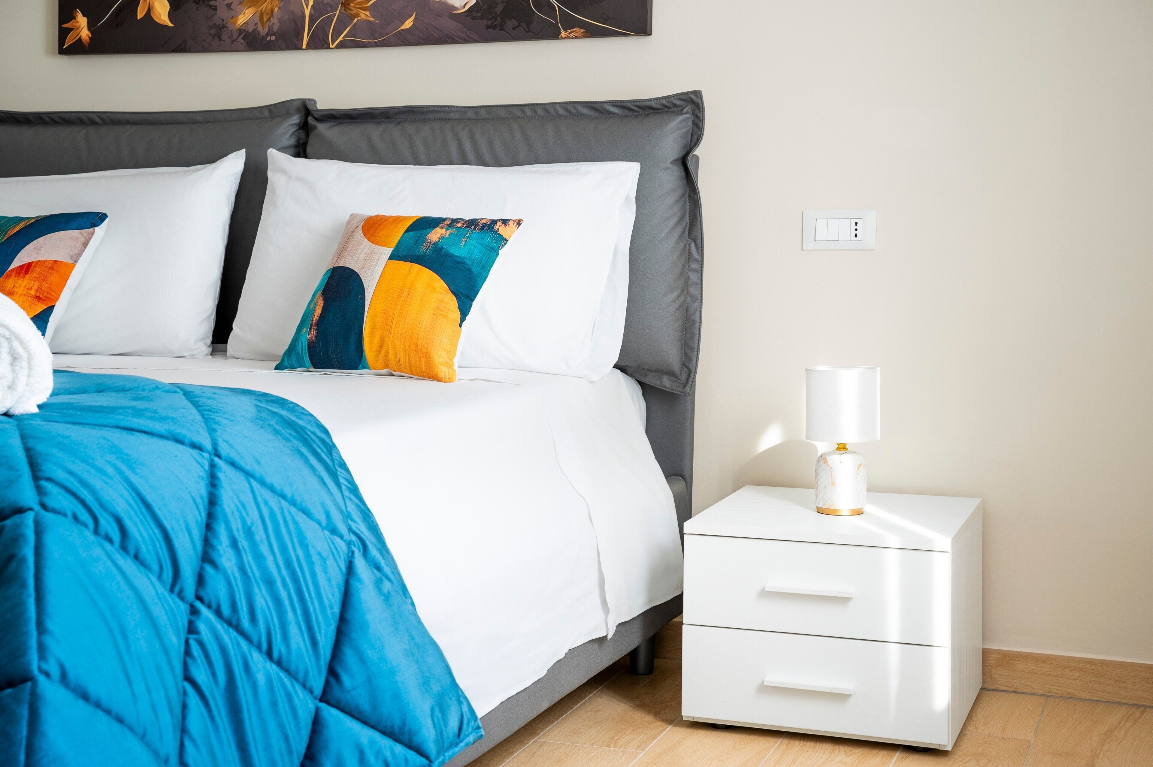Una cama bien tendida con una colcha azul, almohadas blancas y una mesita de noche con una lámpara.