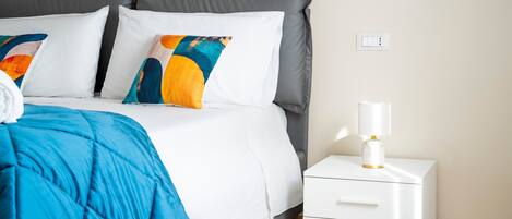 Una cama bien tendida con una colcha azul, almohadas blancas y una mesita de noche con una lámpara.