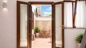 Une pièce dotée d’une porte-fenêtre coulissante donnant sur une terrasse extérieure avec une table et des chaises, un pot de fleurs et un mur en pierre.